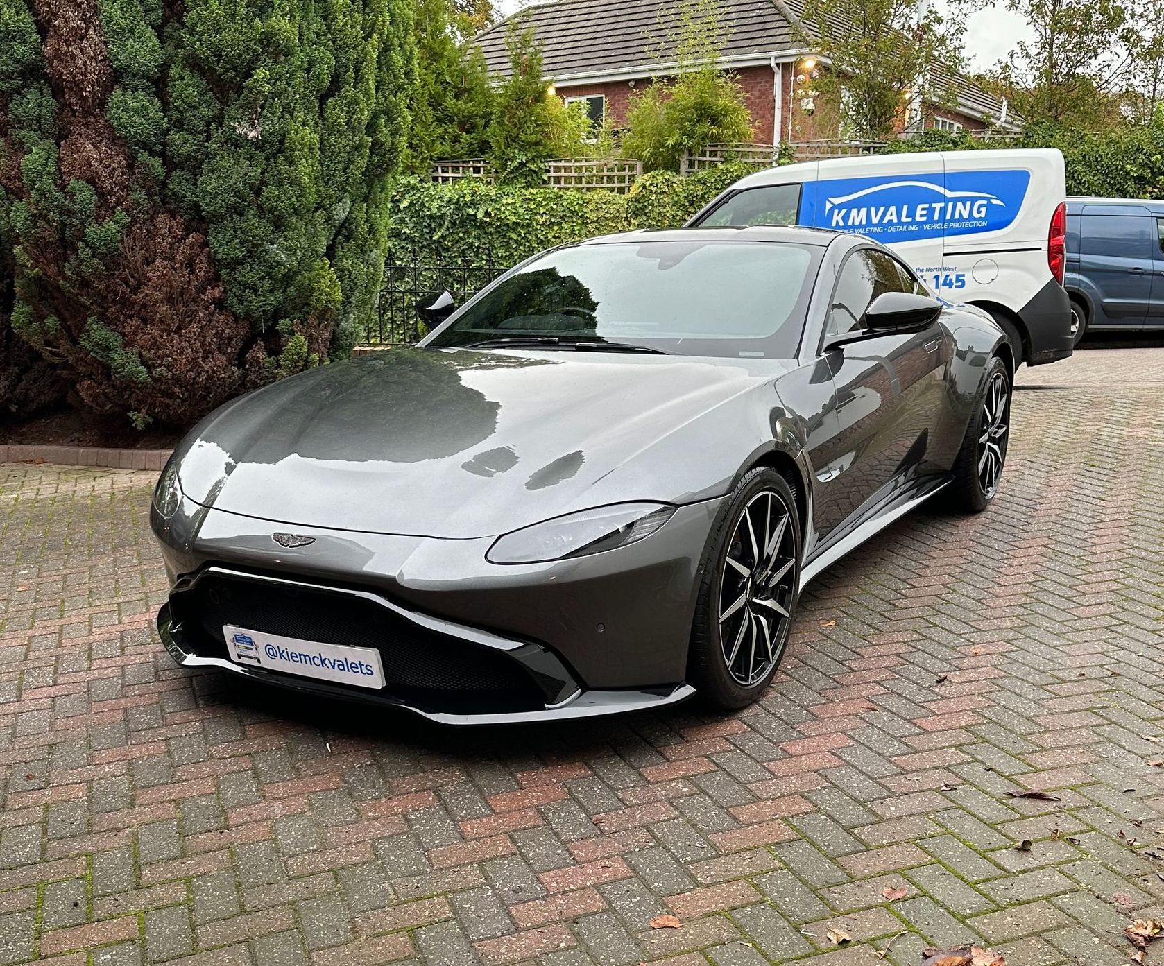 A gray sports car is parked on a brick driveway next to a white van.