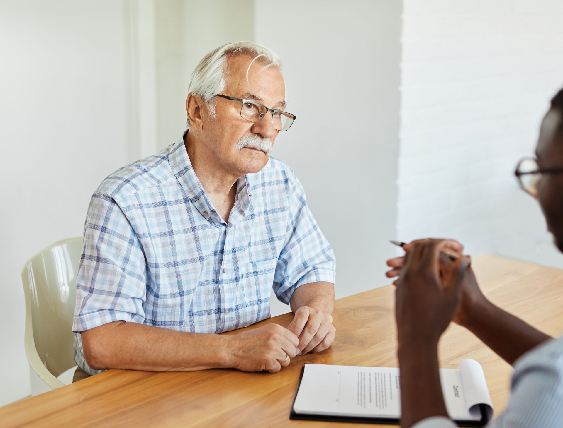Older person with glasses and a plaid shirt speaking to someone at a table, holding a pen and notepad.