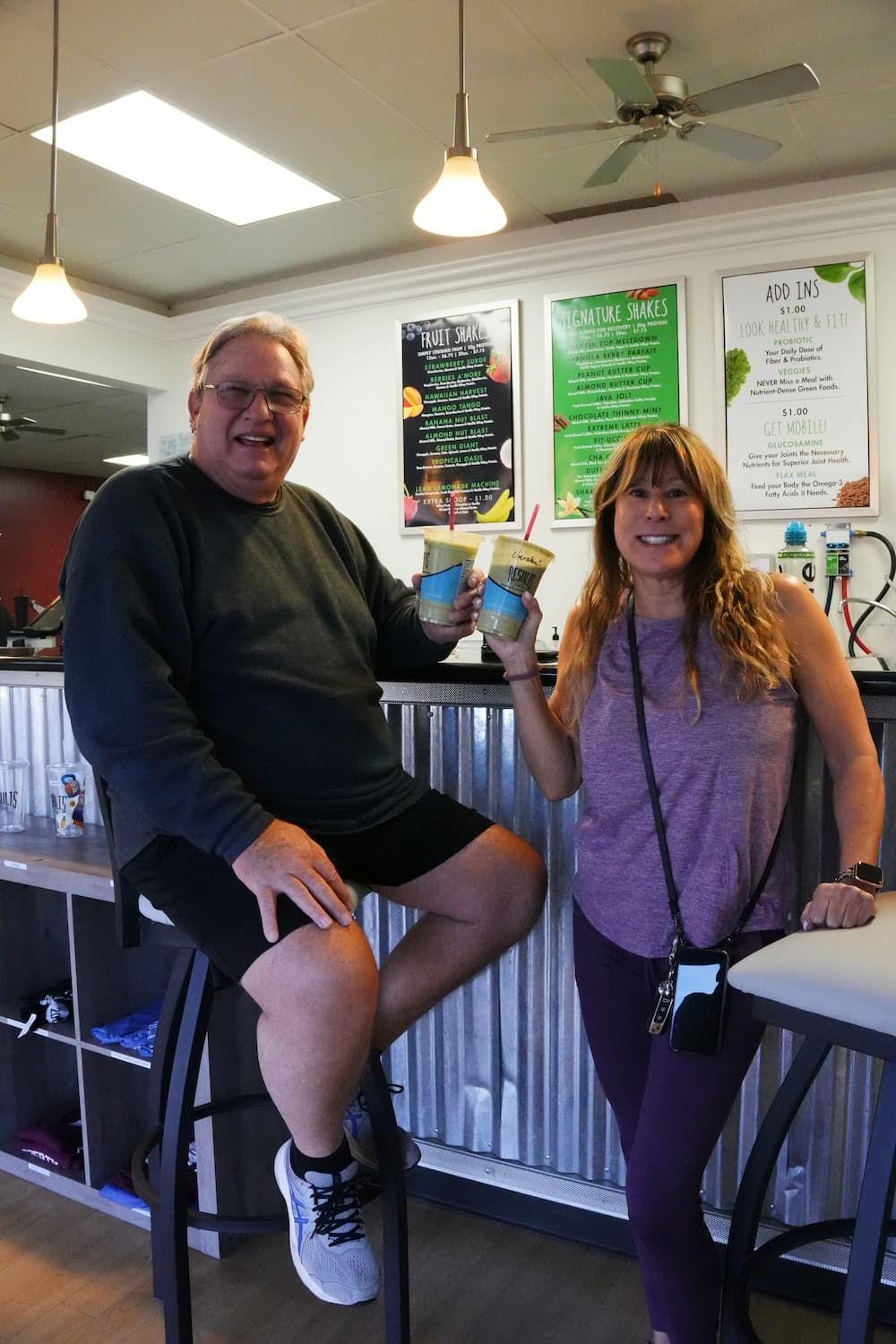 Man and woman in a juice bar, toasting blue-green drinks. Man sits on a stool, woman stands smiling.