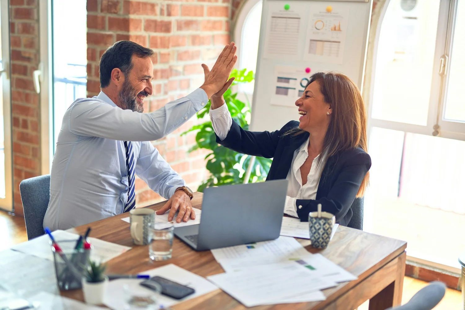 Two coworkers high-five across a laptop at a sunlit office desk