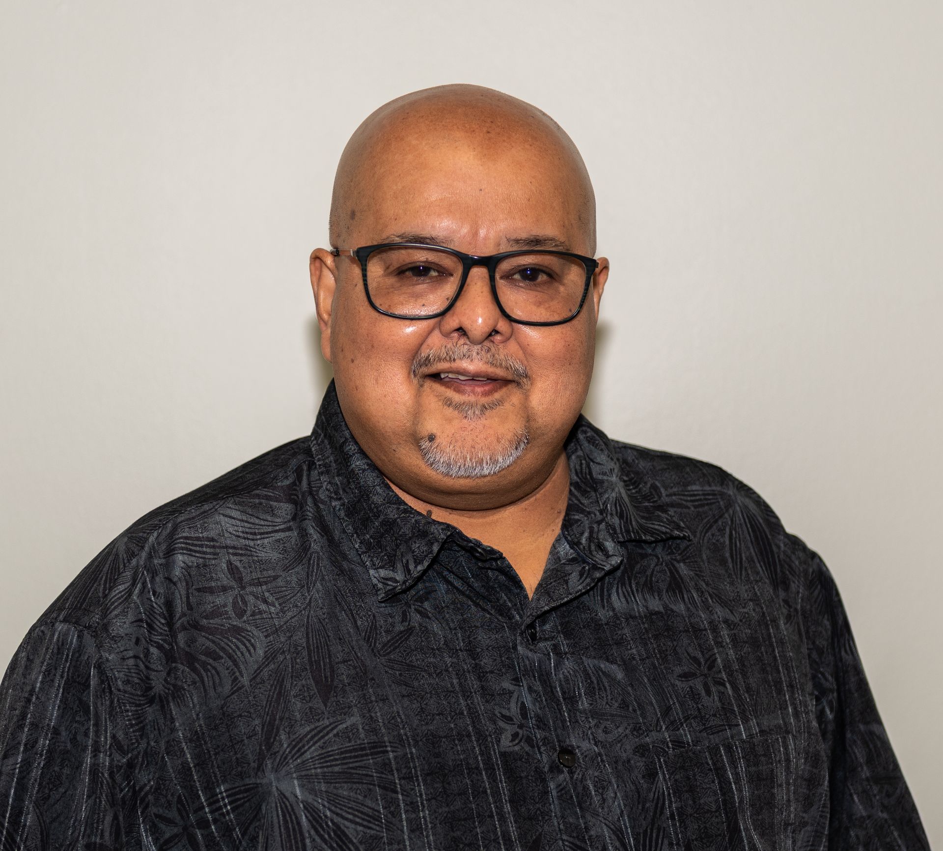 A bald man wearing glasses and a black shirt is standing in front of a white wall.