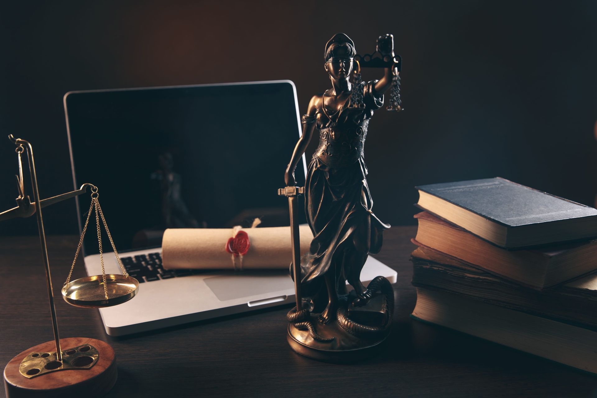 Justice statue beside a laptop, scales, and rolled legal document on a dark desk