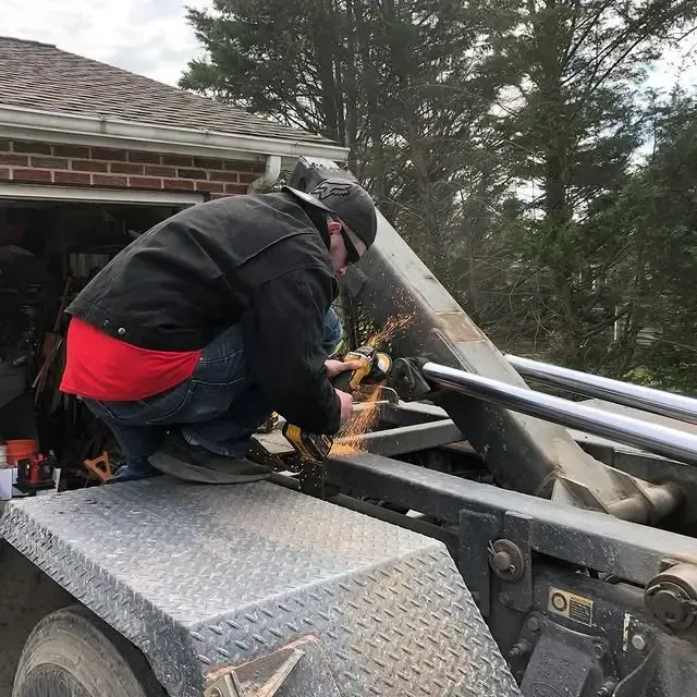 A man is kneeling on the back of a dump truck while using a grinder.