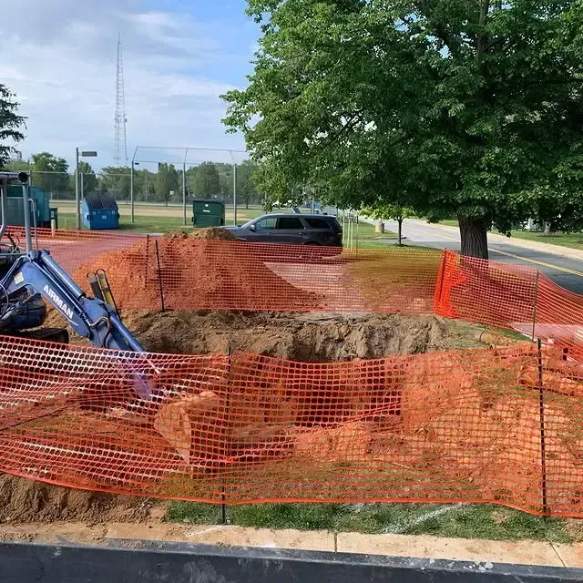 A blue excavator is digging a hole in the ground behind an orange fence.