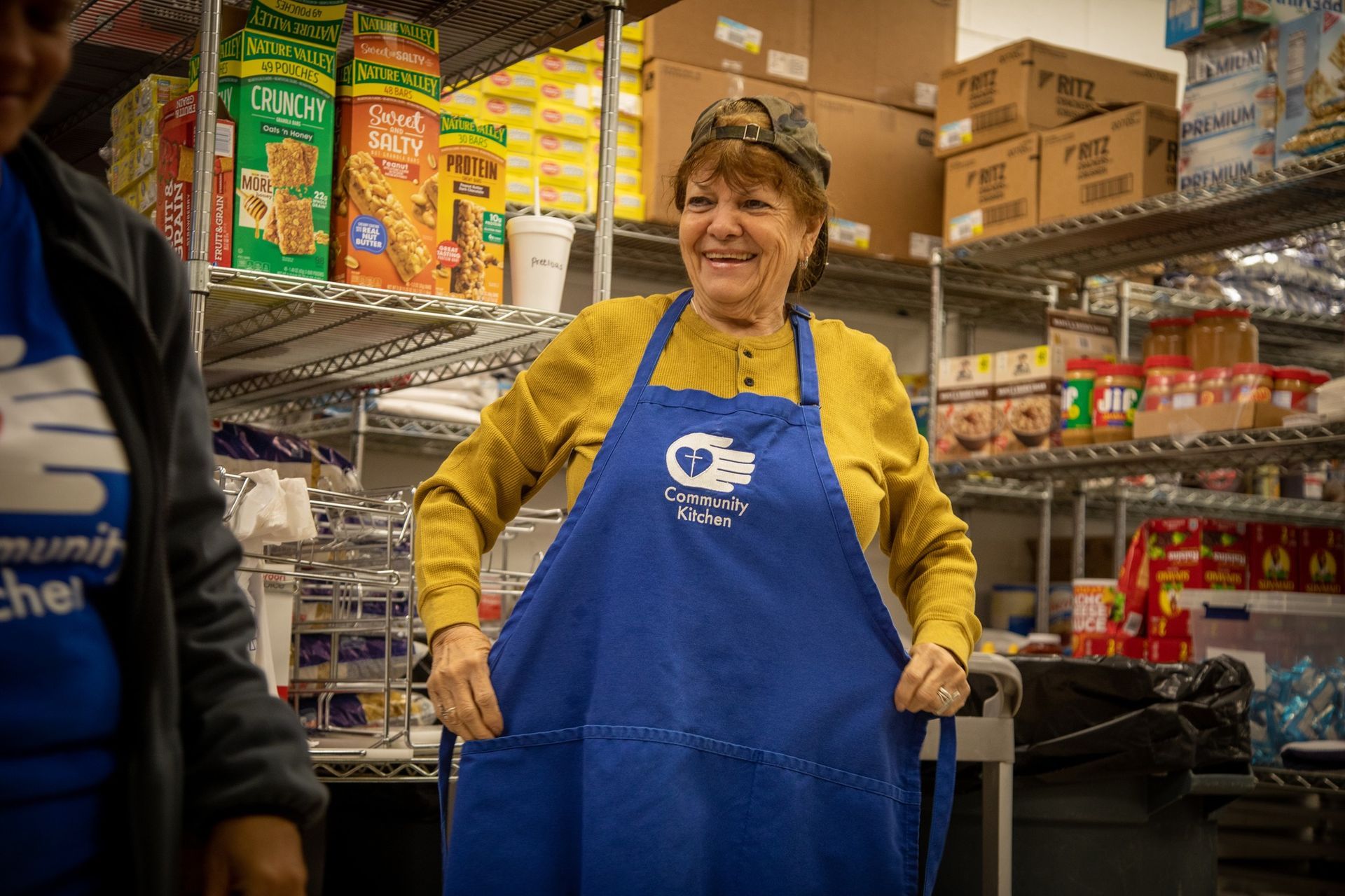 Woman wearing a blue apron smiles in a pantry. Shelves hold food boxes.