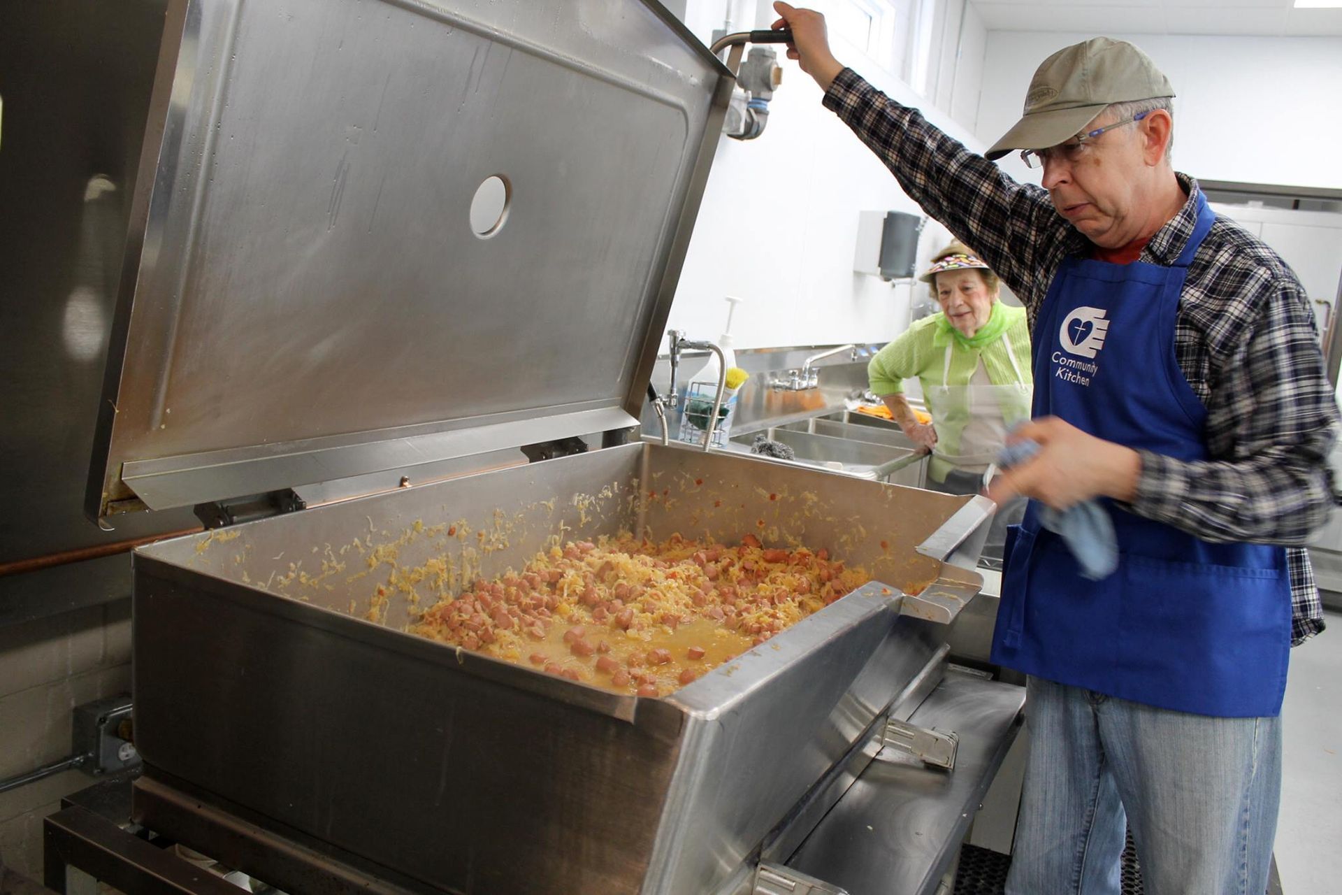 Man stirring large pot of food with woman watching in commercial kitchen.