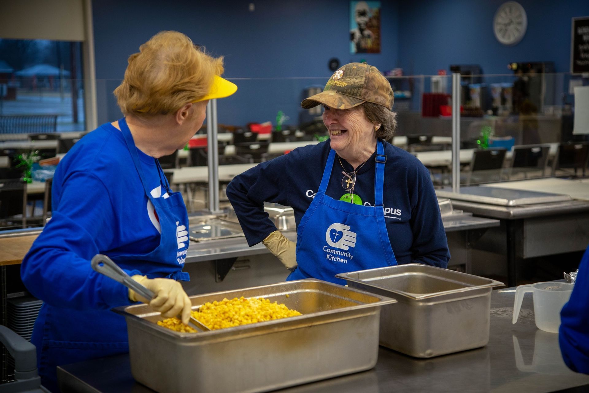 Two women in blue aprons serve food in a kitchen. One stirs a yellow dish; both smile.