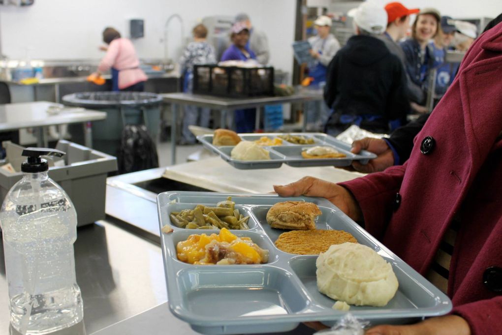 Person holding cafeteria trays with food, others line up; kitchen background.