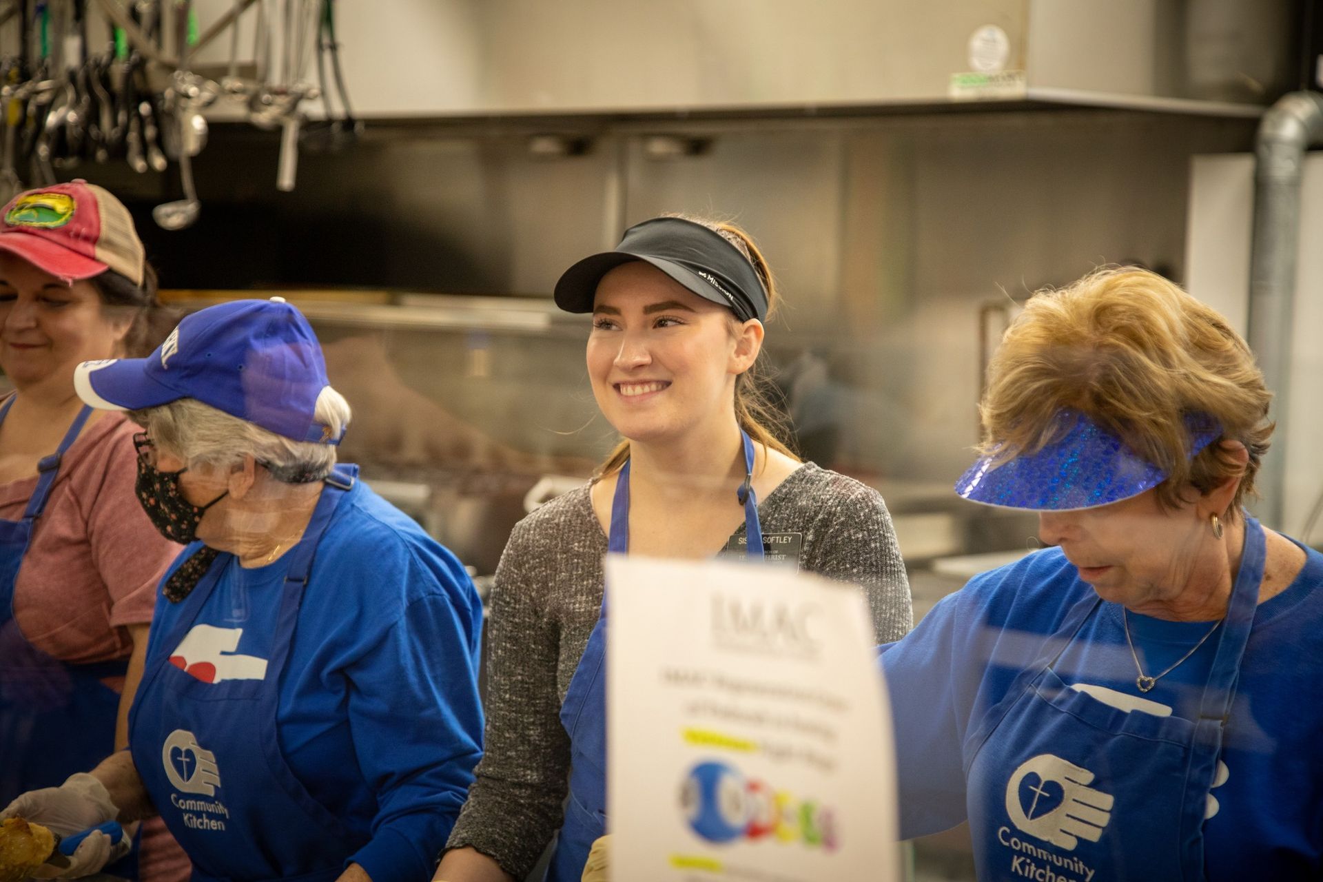 People in a kitchen wearing blue aprons volunteering. A young woman smiles, wearing a visor.