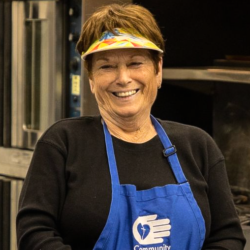 Woman in blue apron smiles, wearing visor, in a kitchen.