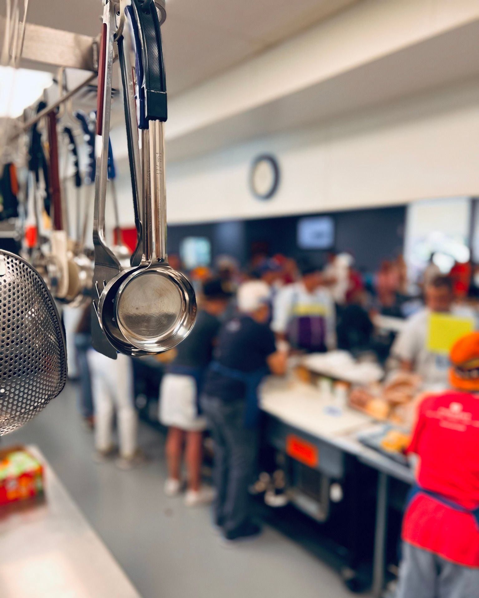 Kitchen utensils hanging, people serving food at a counter in a cafeteria.