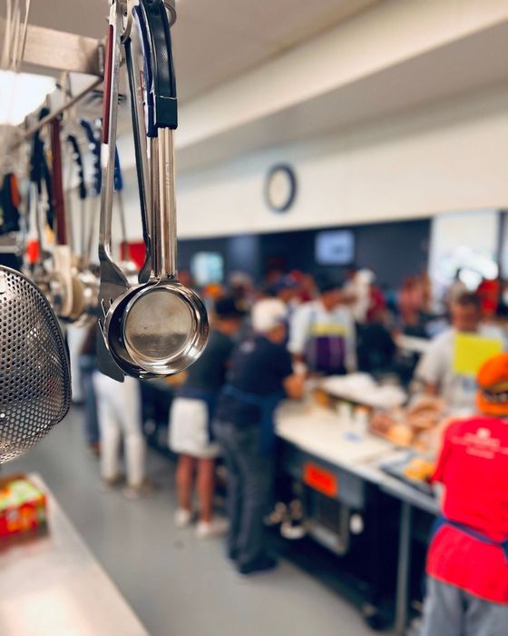 Kitchen utensils hanging, people serving food at a counter in a cafeteria.