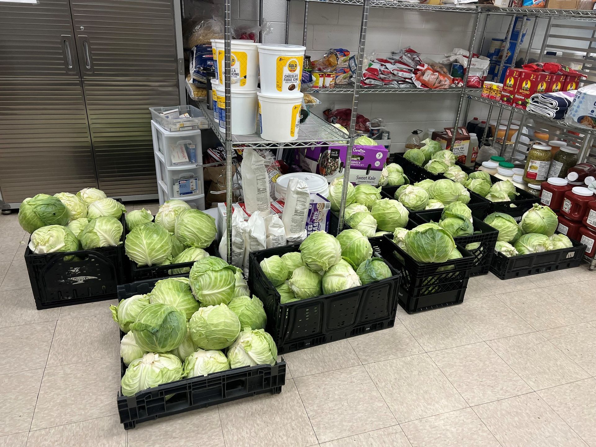 Crates of green cabbages in a storage room, next to shelves with food items.