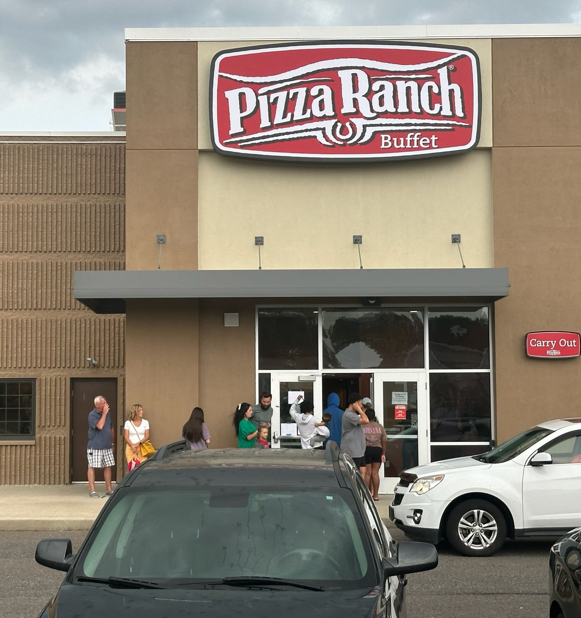 Pizza Ranch Buffet entrance; people line up outside under a gray awning, cars in foreground.