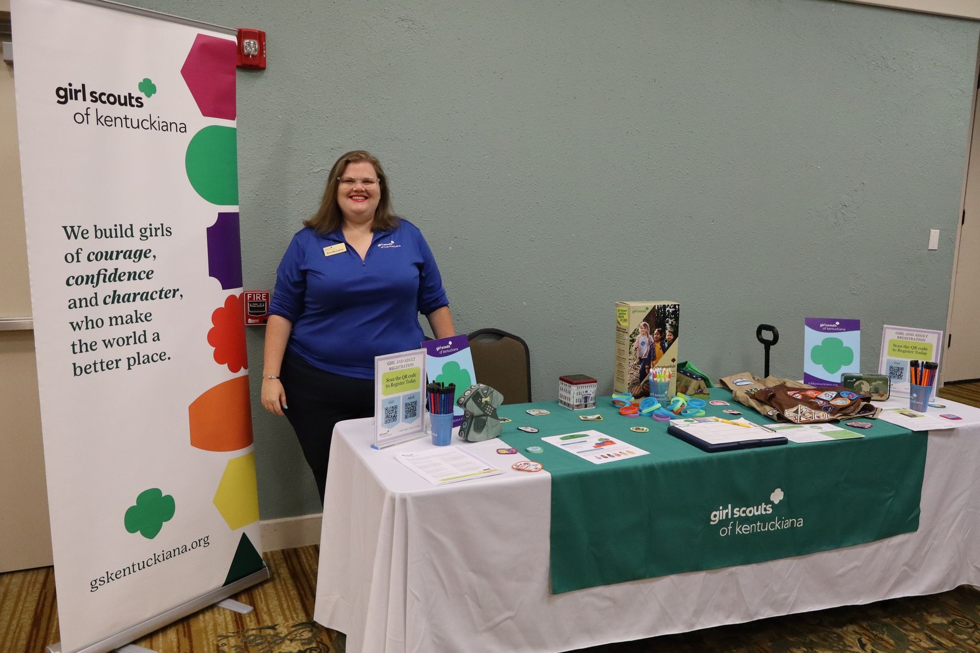 Woman at a Girl Scout booth with table display and banner, wearing a blue shirt, smiling.