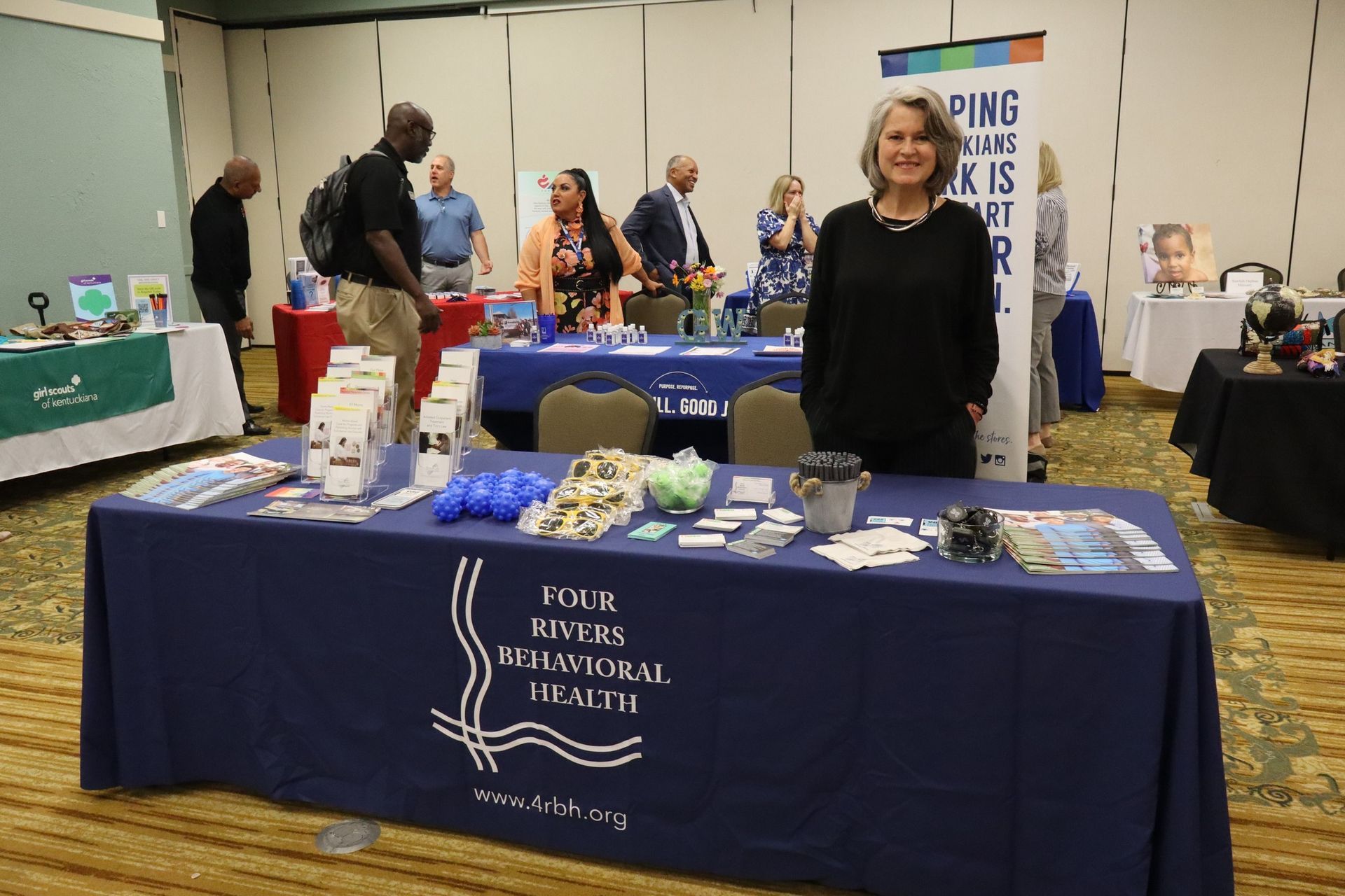 Woman stands at a booth for Four Rivers Behavioral Health, with brochures and swag on display at an event.
