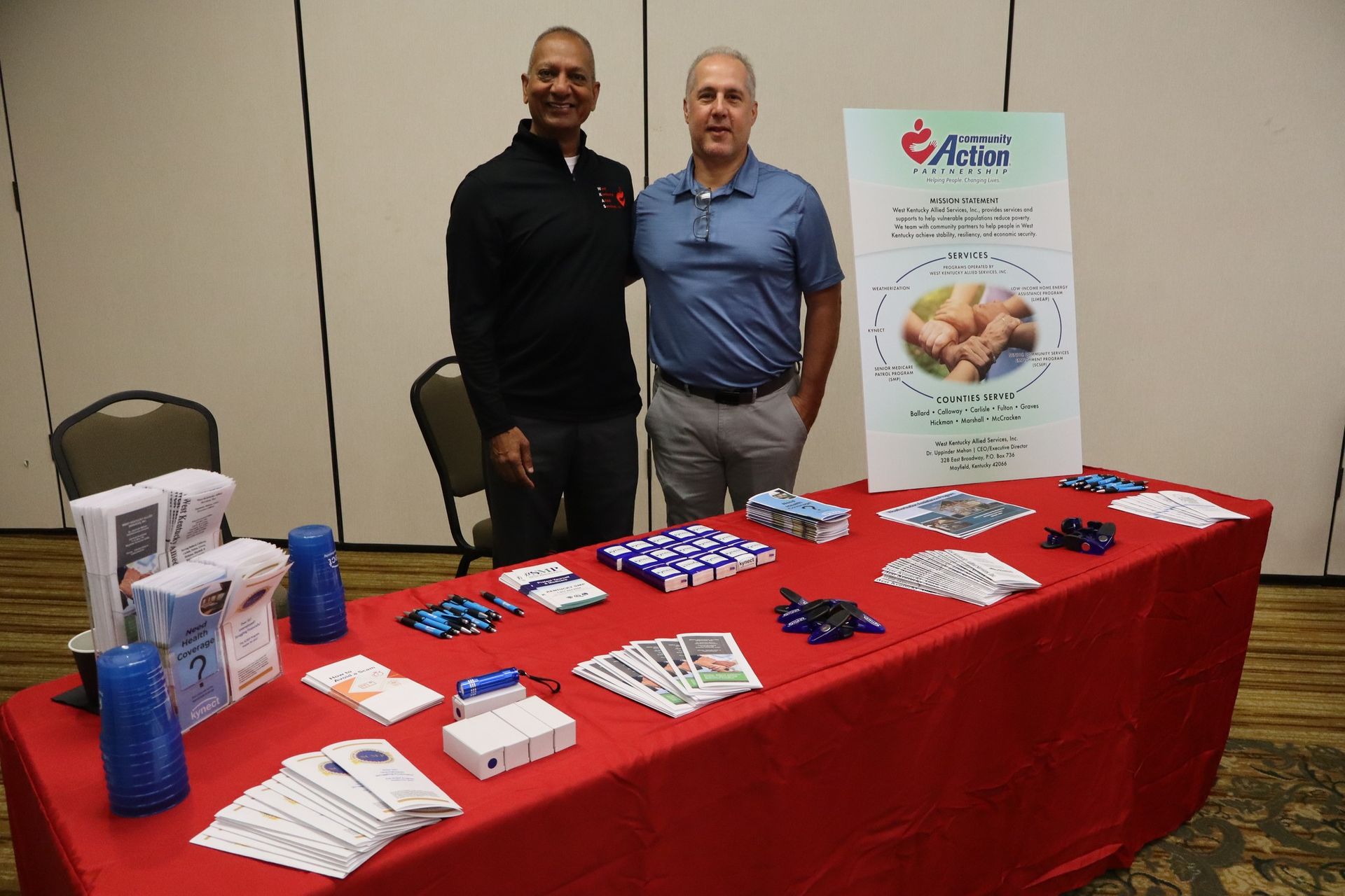 Two men stand behind a table displaying medical supplies. Red tablecloth, sign with product images.