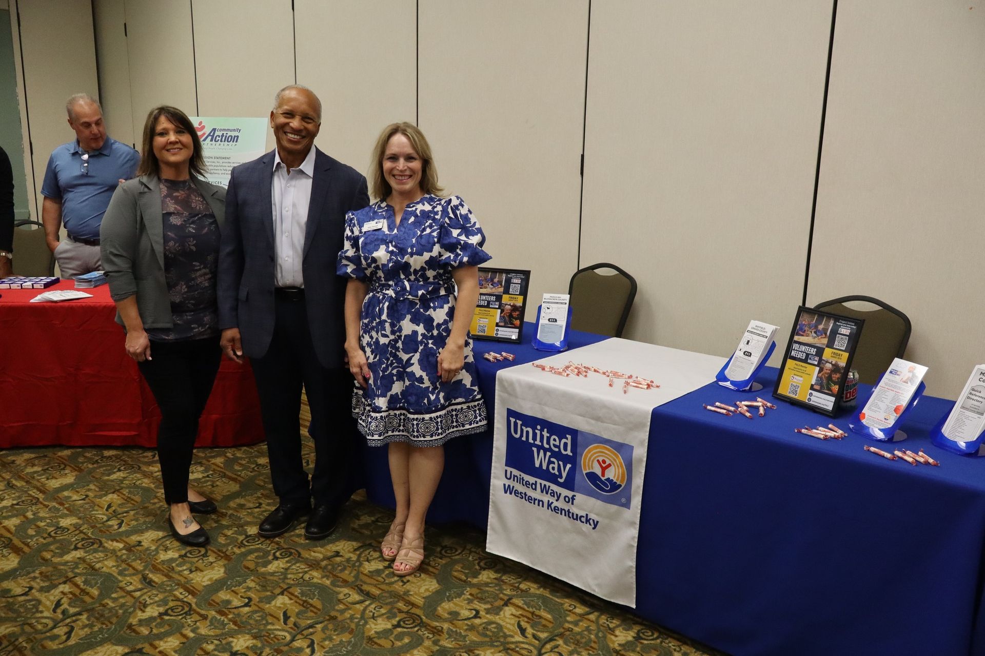 Three people stand by United Way table at an event. Woman in blue dress smiles, two others look toward camera.