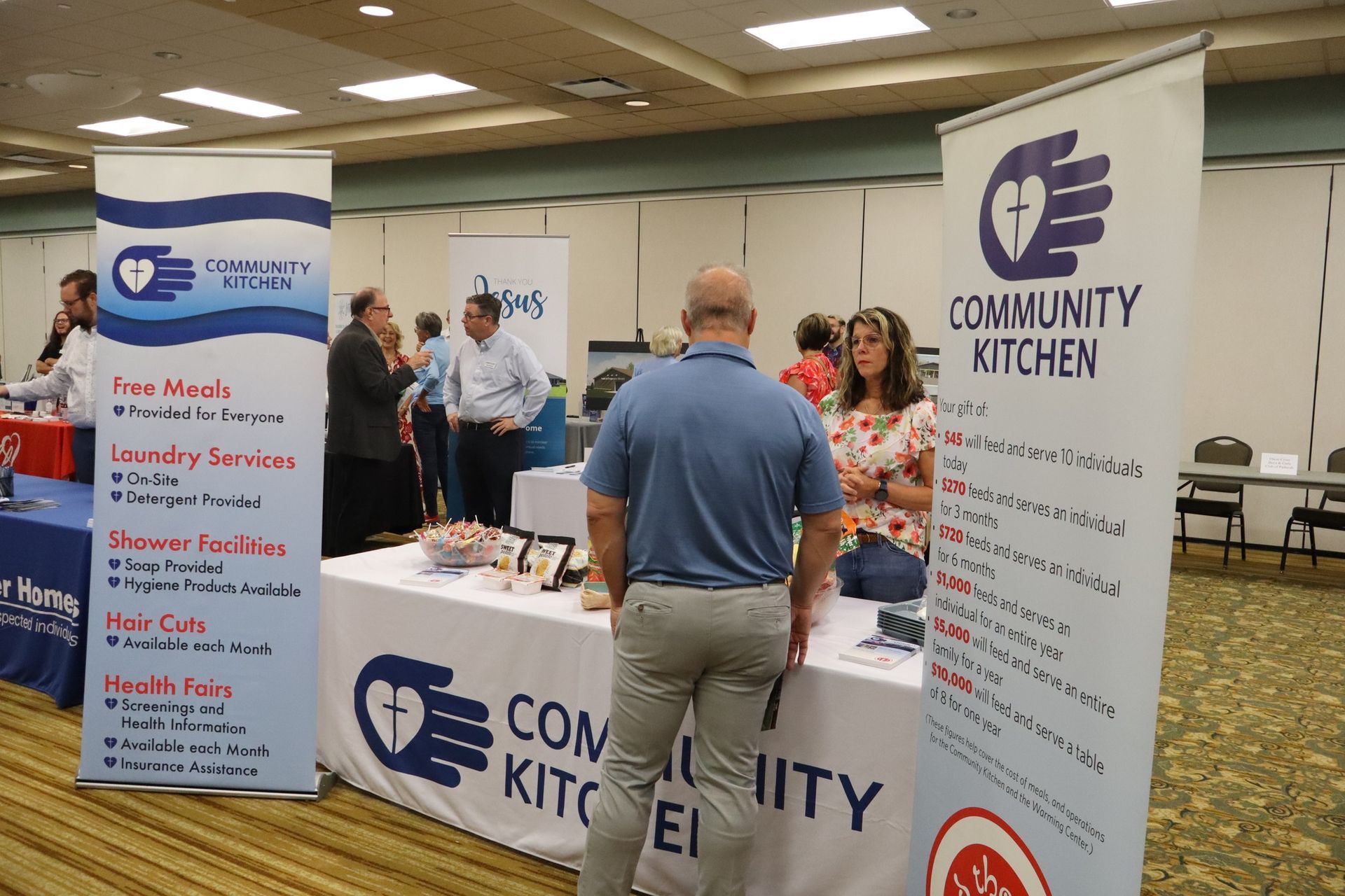 People at a Community Kitchen event. Booth with blue and white banners, people talking.