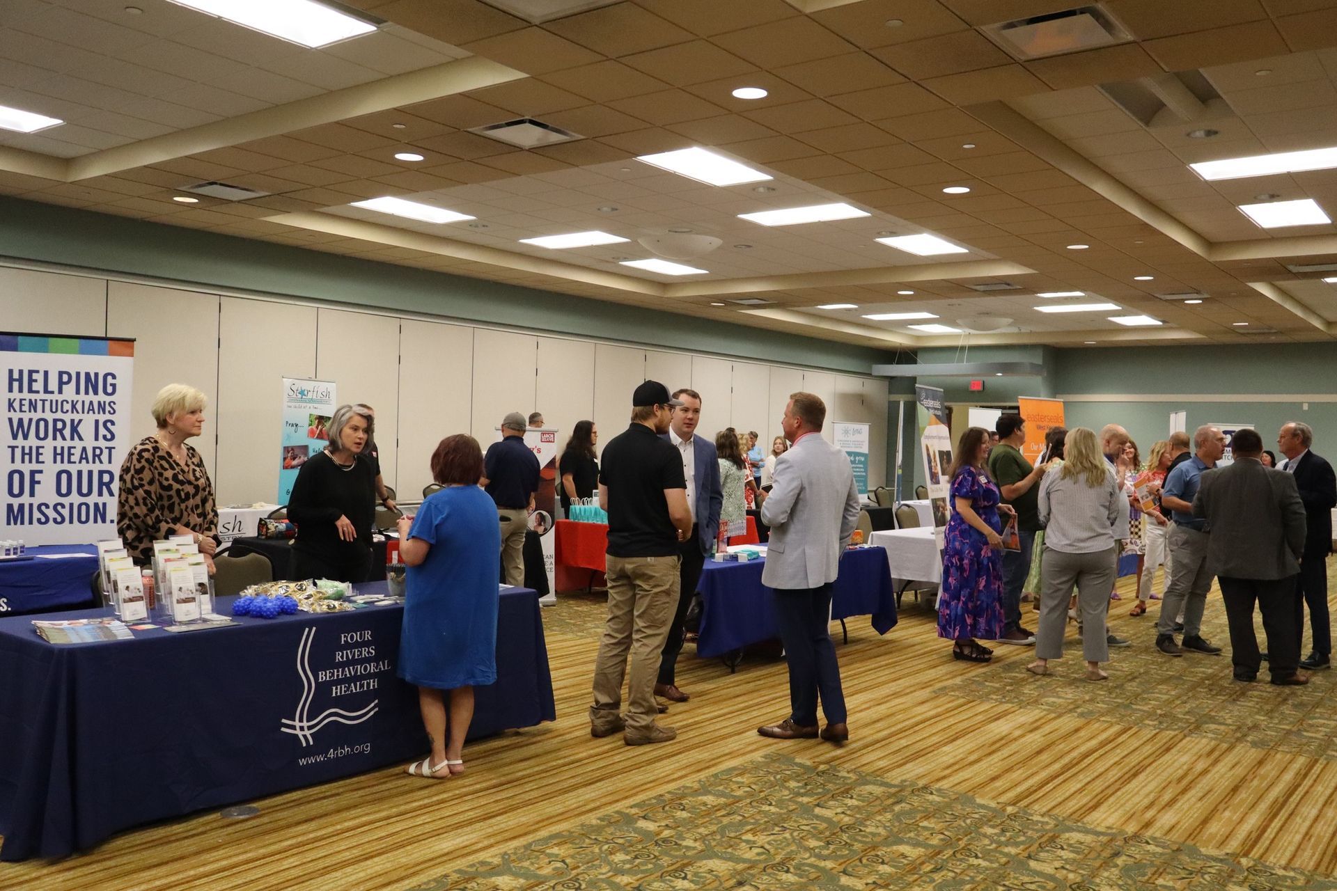 People at an indoor event, possibly a fair or conference, with booths and tables set up.