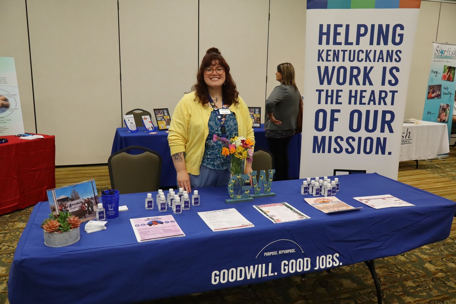 Woman at a Goodwill booth smiling; blue table with products and information, a sign behind her.