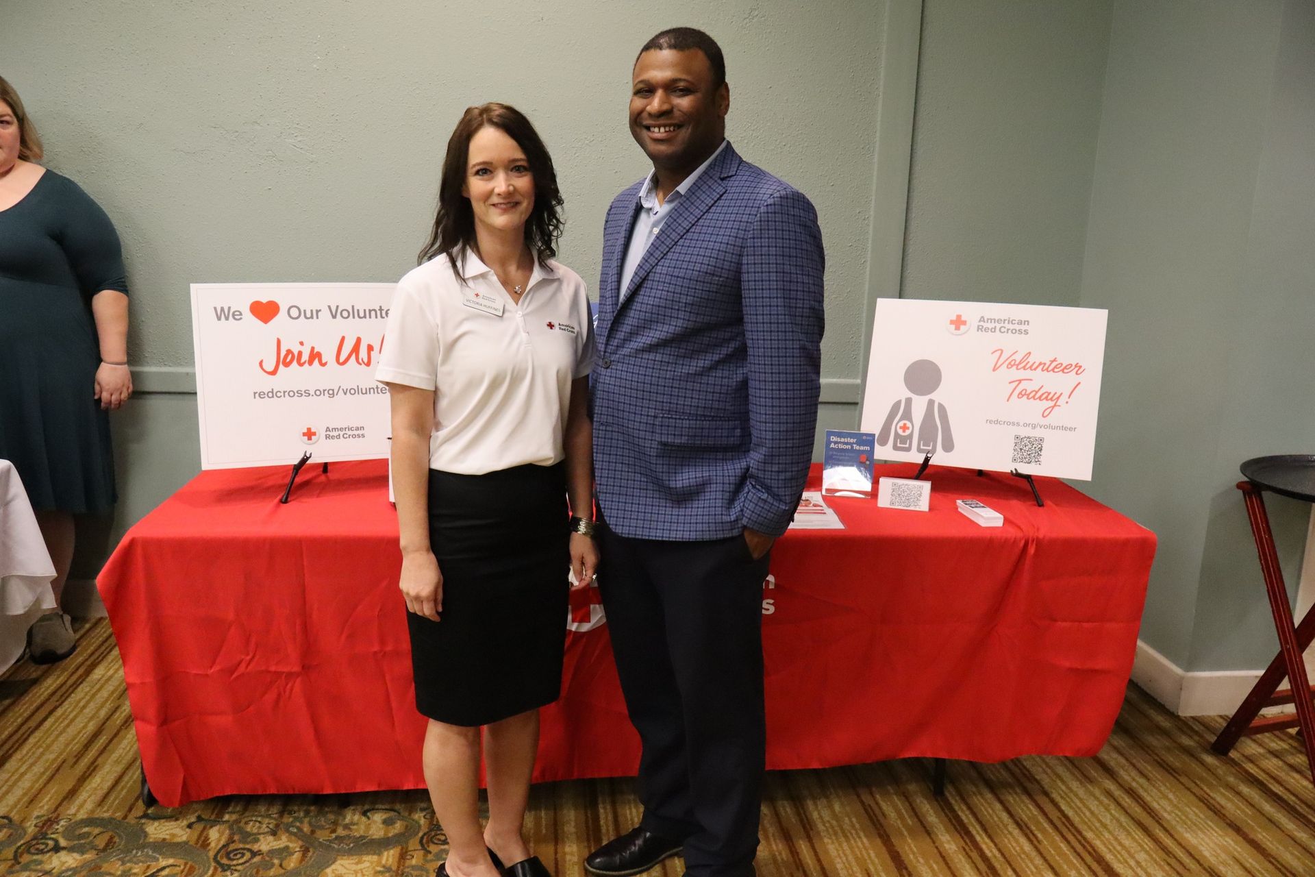 Woman and man standing in front of a red table with signs. Woman wears a white polo shirt and black skirt. Man wears a blue blazer.