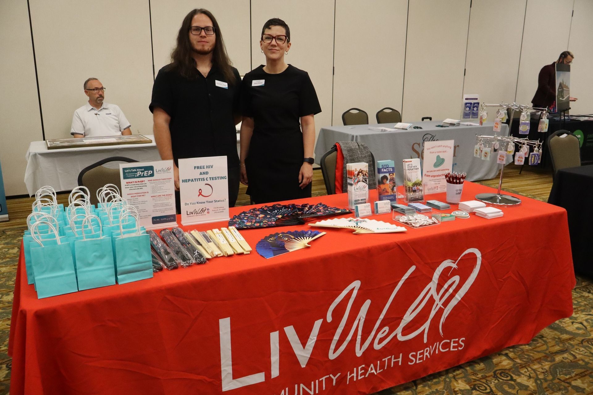 Two people at a LivWell Community Health Services booth with brochures. Red table cloth, light blue gift bags.