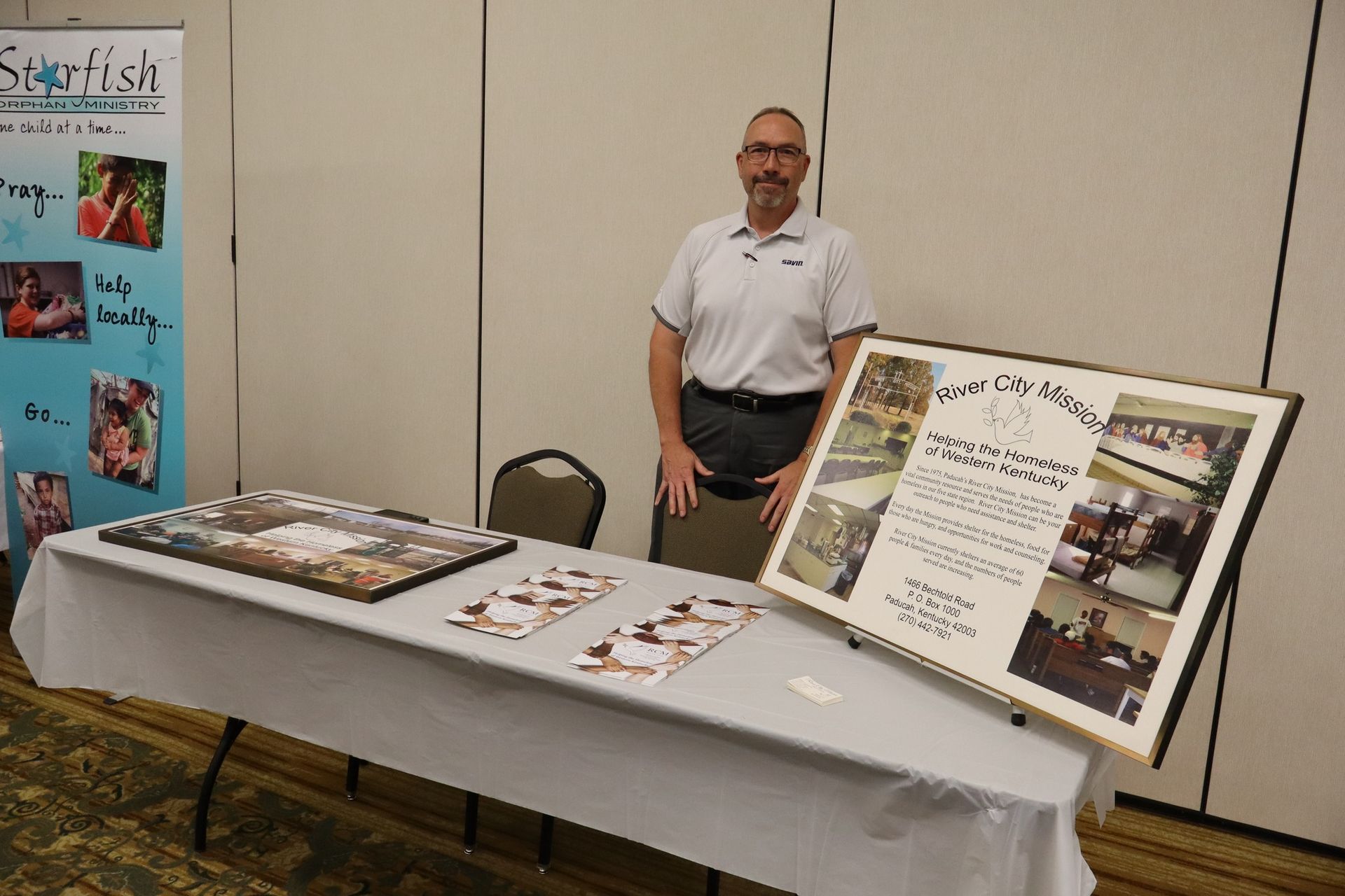 Man stands at table with framed photos and a banner in a conference room.