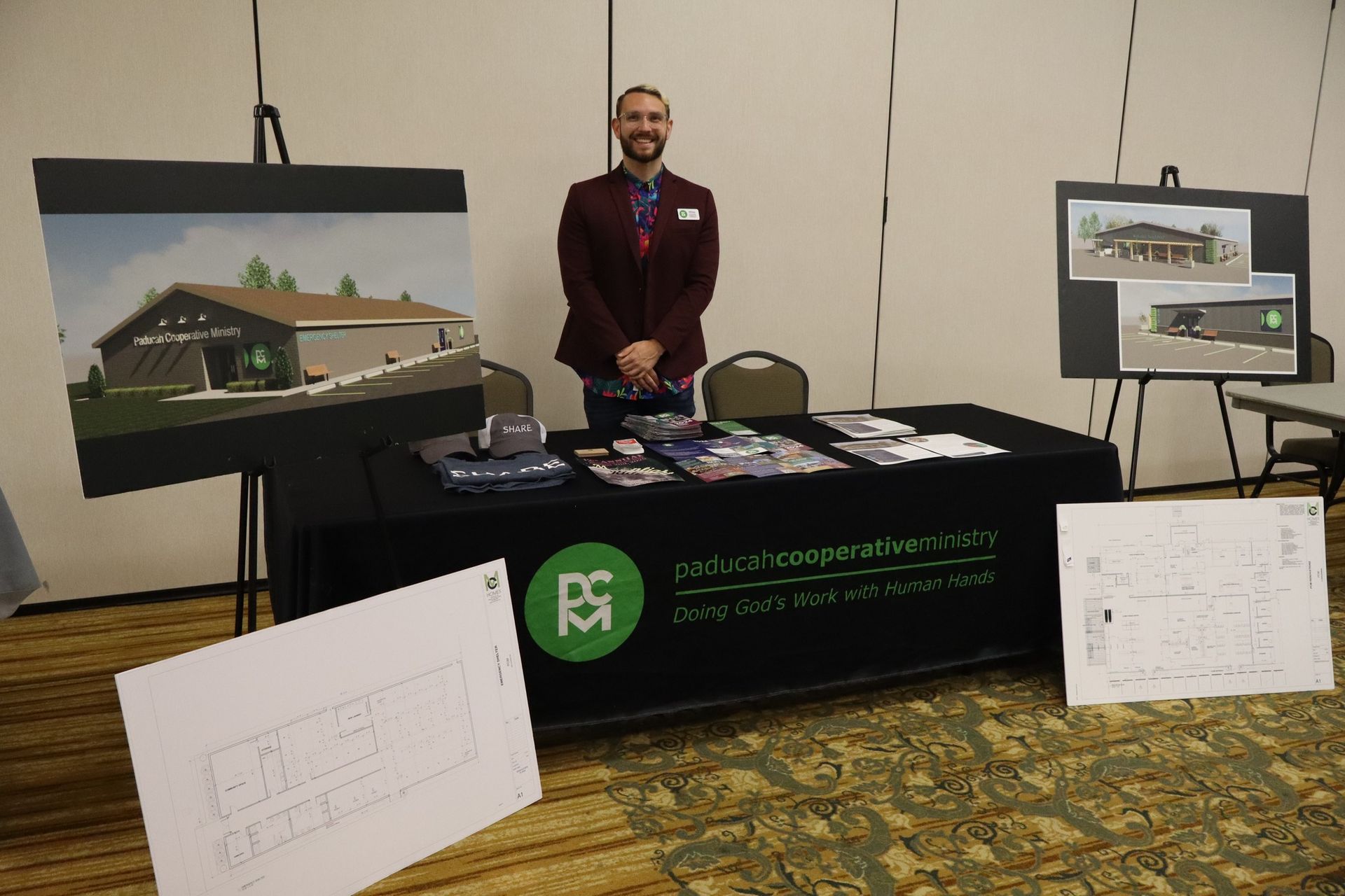 Man stands at a booth with architectural renderings and brochures, promoting a business.