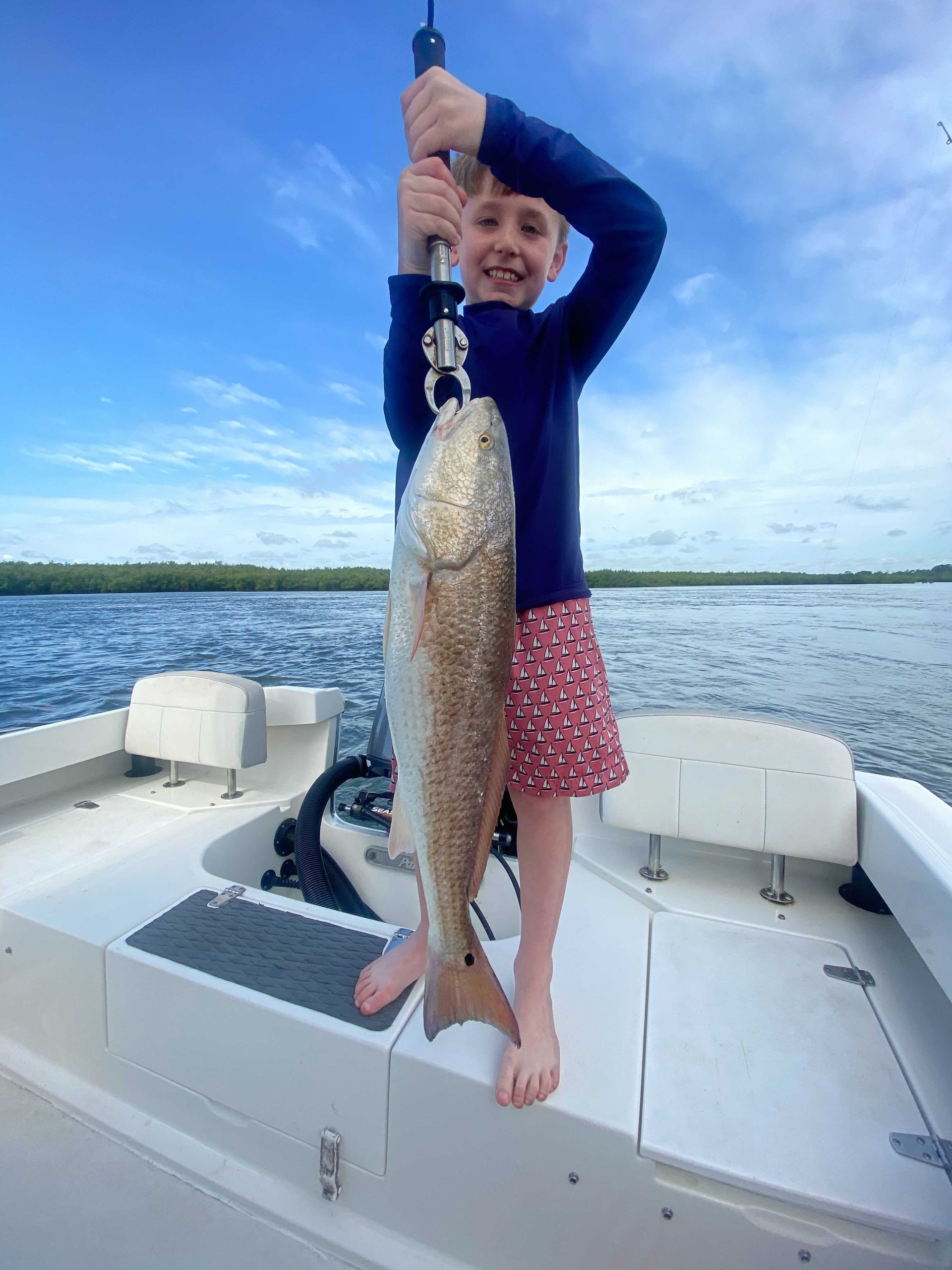 Young child on a boat, holding up a large redfish with a wide smile. Outdoors with blue sky and water.