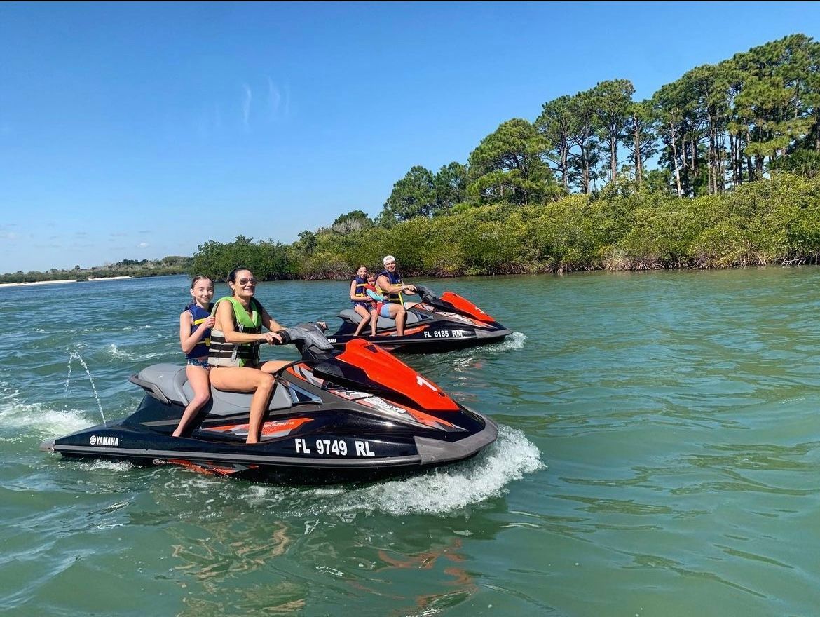 Two jet skis on a waterway with people riding them under a blue sky and greenery.