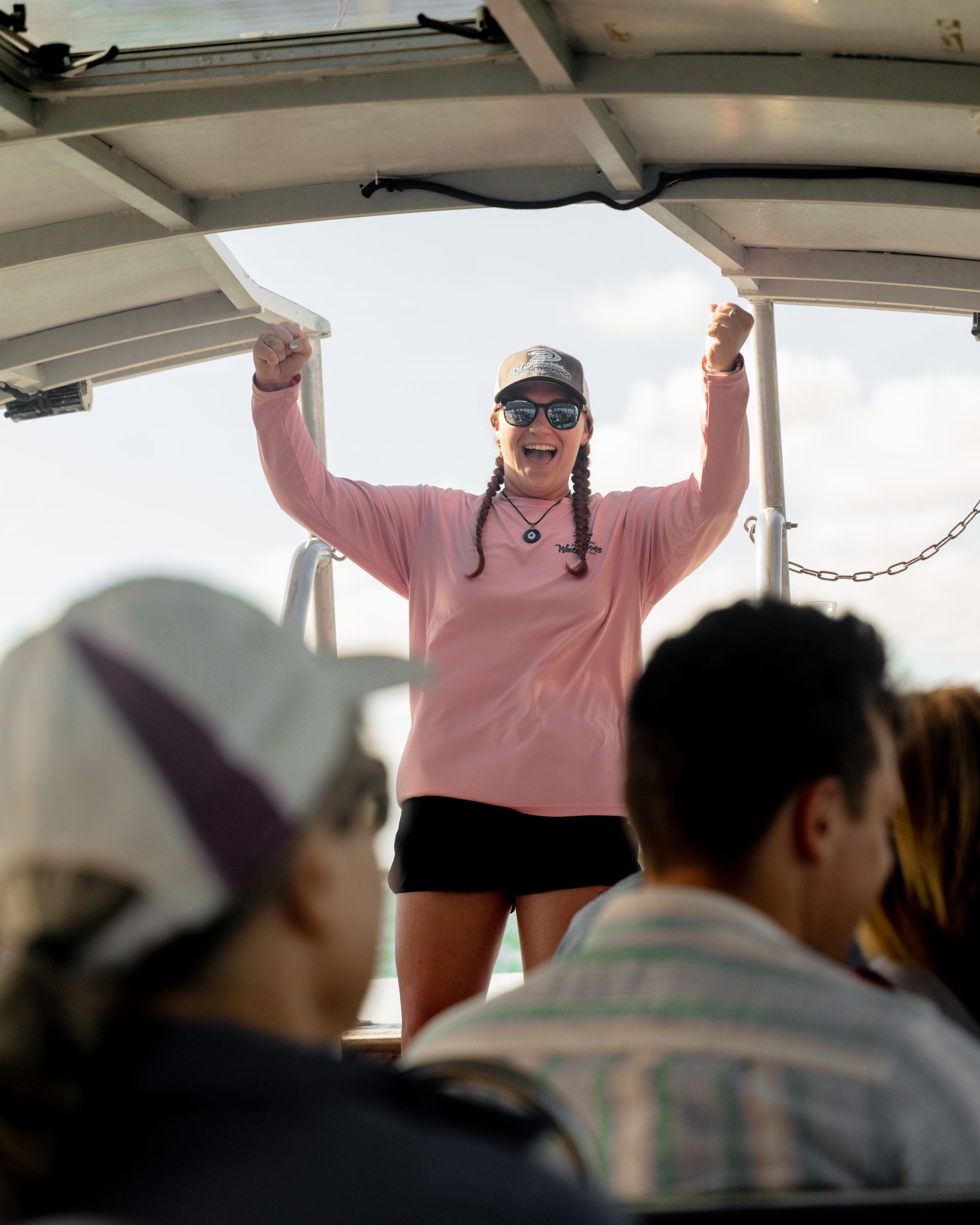 Woman on a boat with arms raised in celebration, wearing pink shirt, black shorts, and hat.