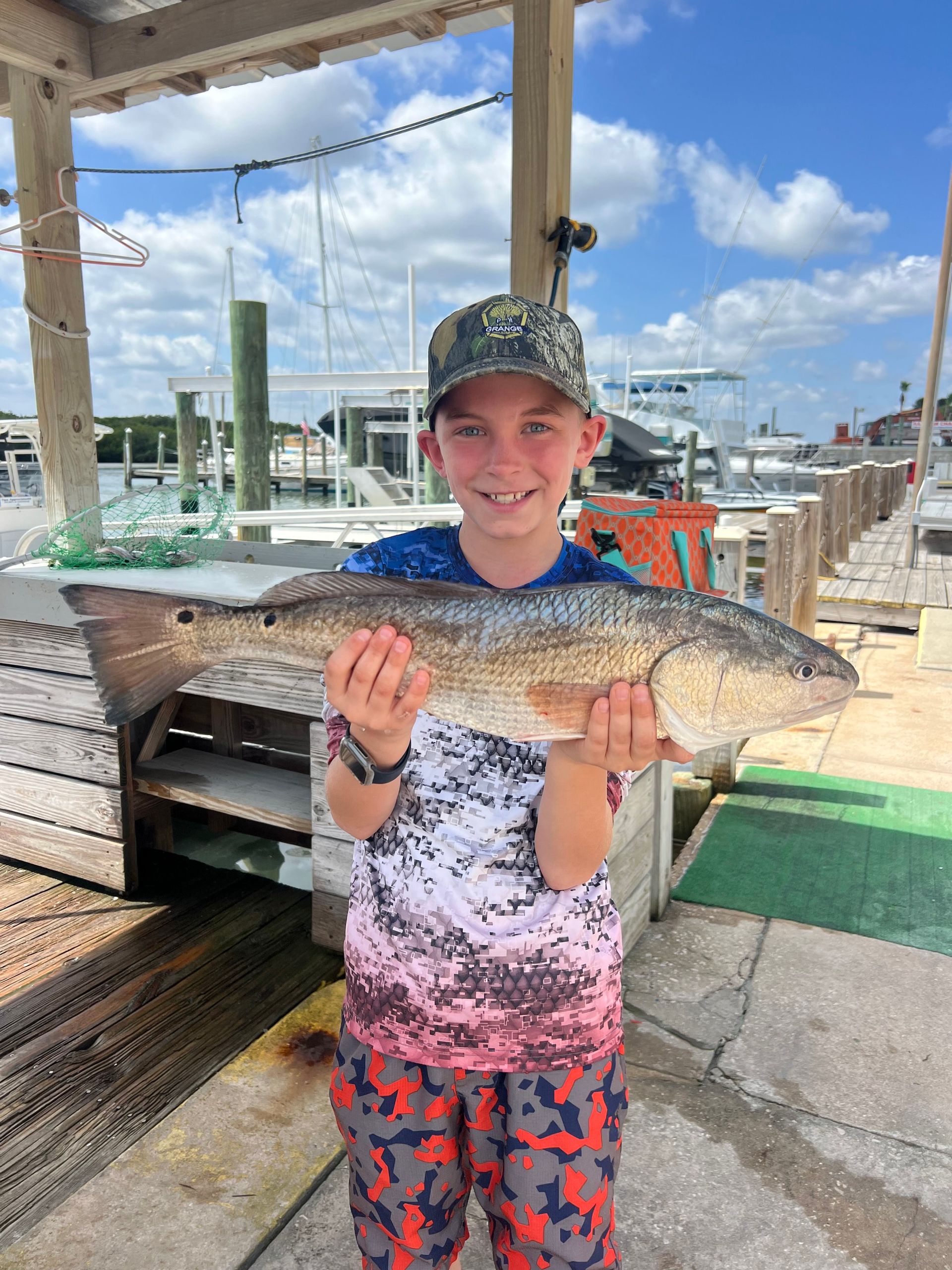 Young boy smiles, holding a large reddish fish on a dock near boats under a bright sky.