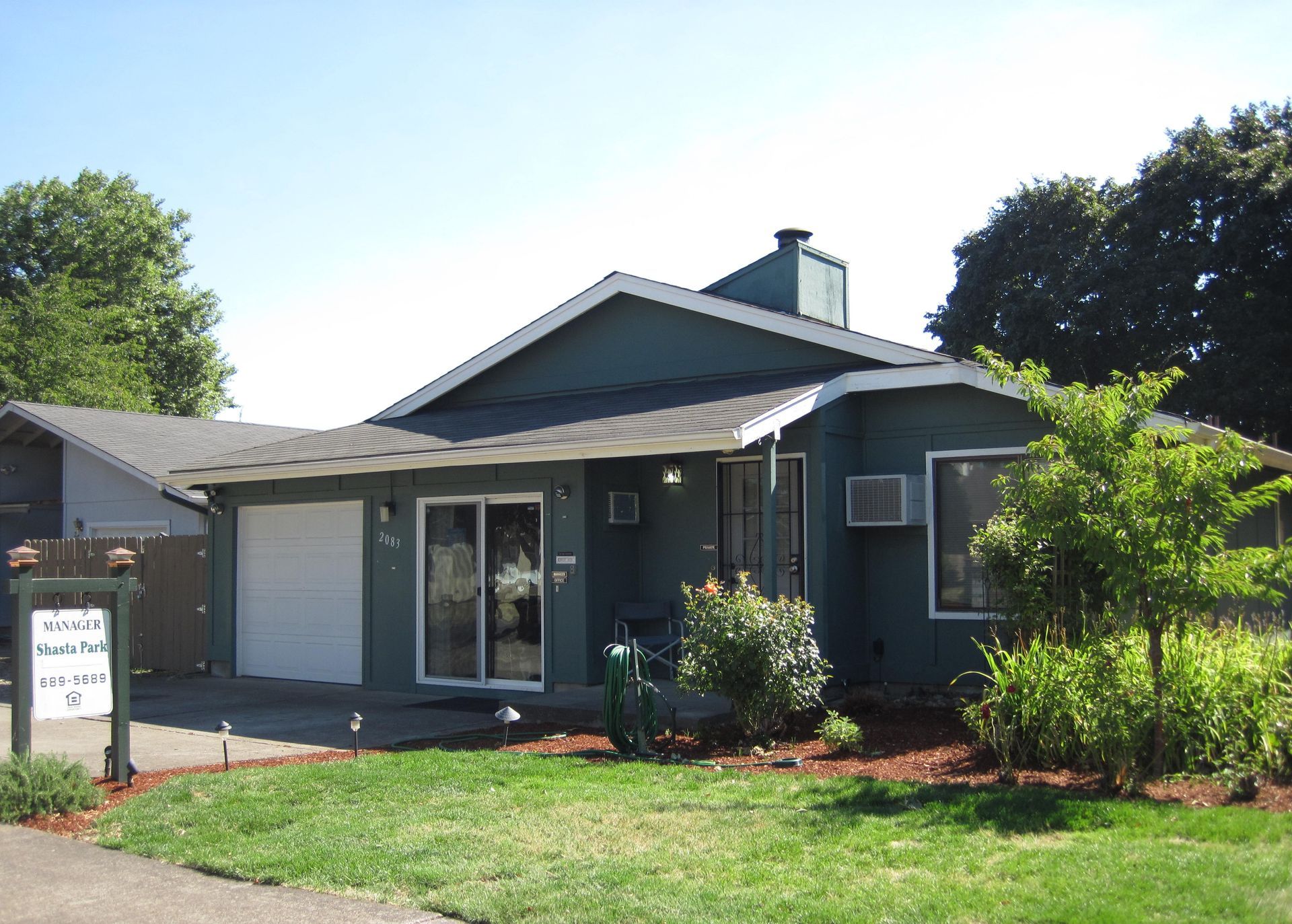 Green house with a garage and sign in front, surrounded by trees and grass.