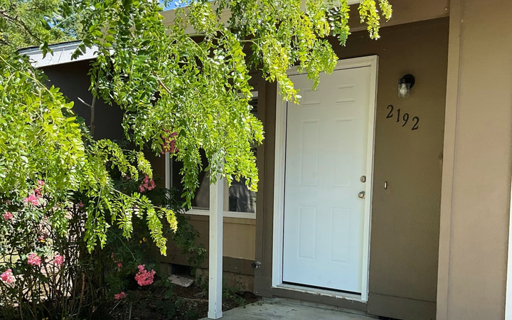 White door of a beige house with the number 2392. Green bush with pink flowers in the foreground.
