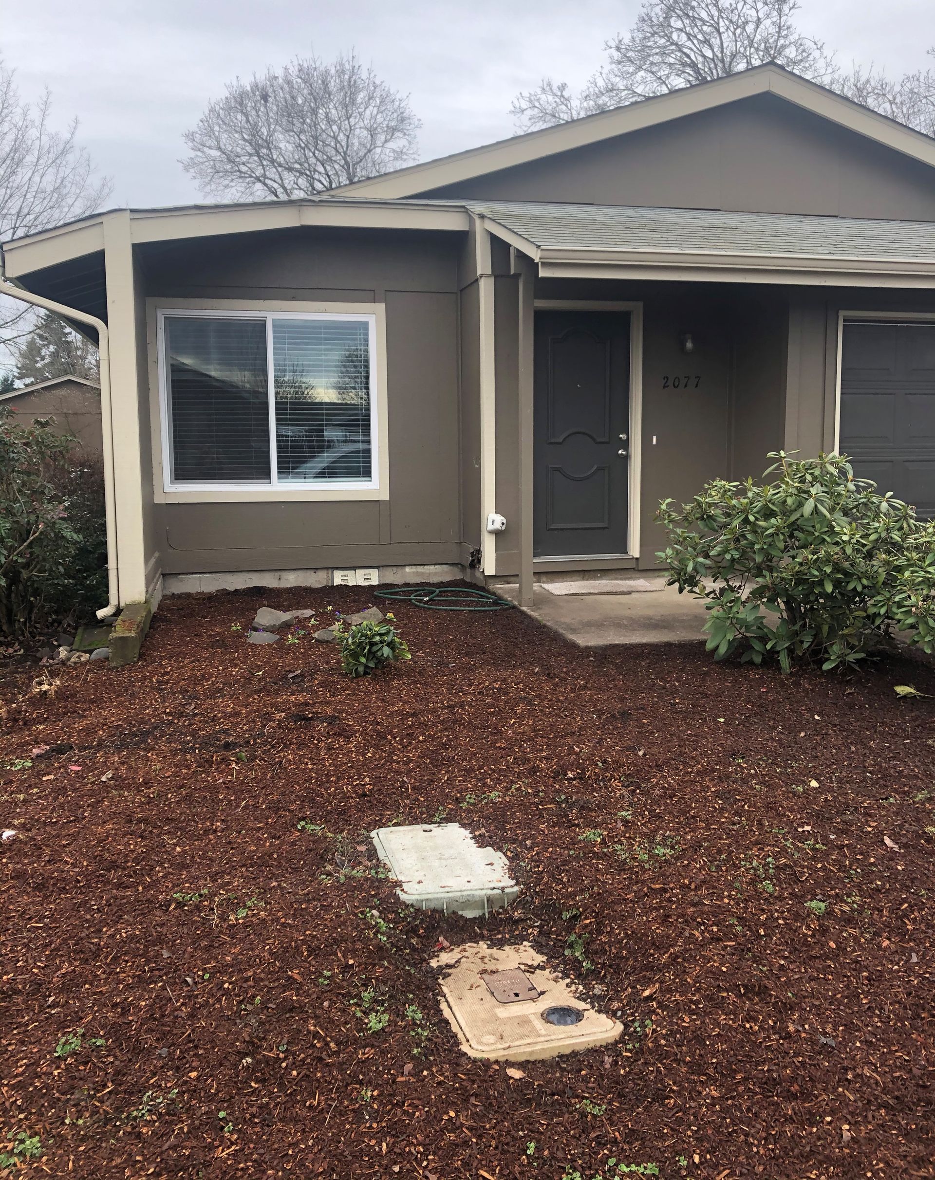 Brown house with mulch landscaping, door, window, and bushes.