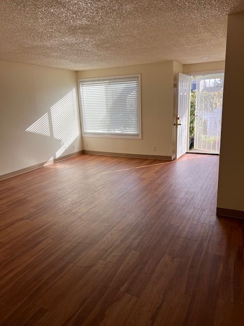 Empty living room with wood flooring, a window with blinds, and a door. Sunlight streams in.