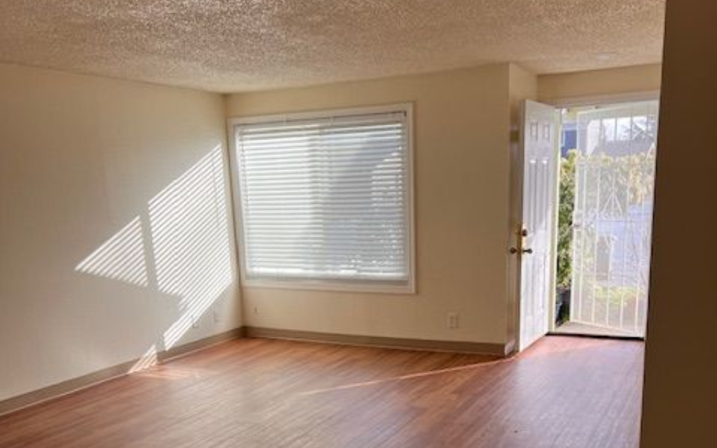 Empty room with wood-look flooring, closed window with blinds, and open door with a gated exterior. Sunlight on wall.