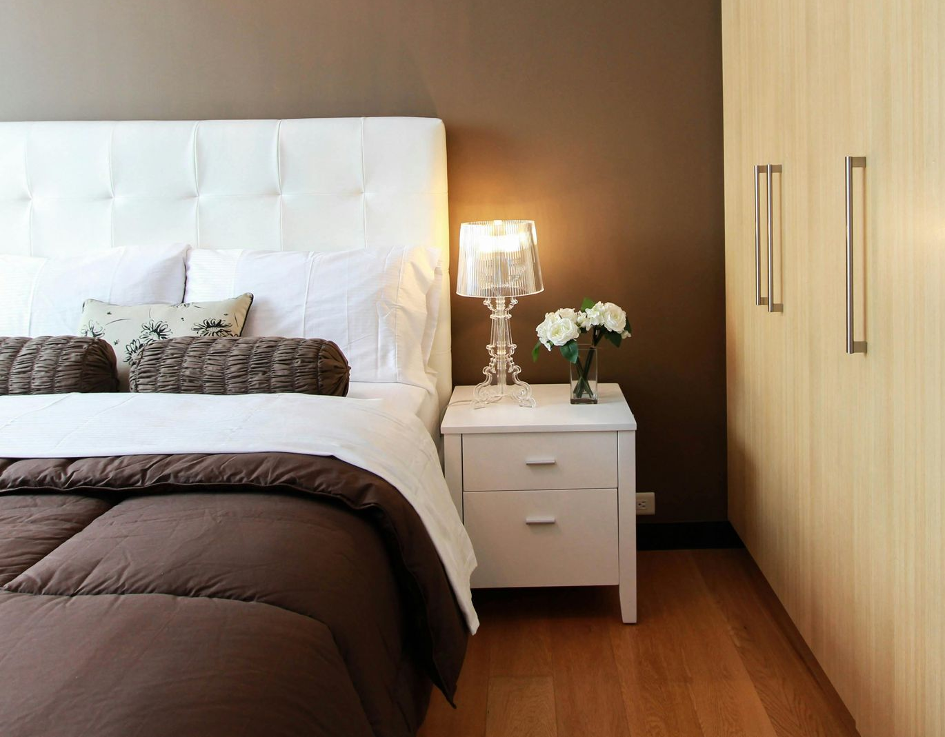 Bedroom with bed, nightstand, wood closet, and full-length mirror reflecting plants. Brown, white and wood colors.