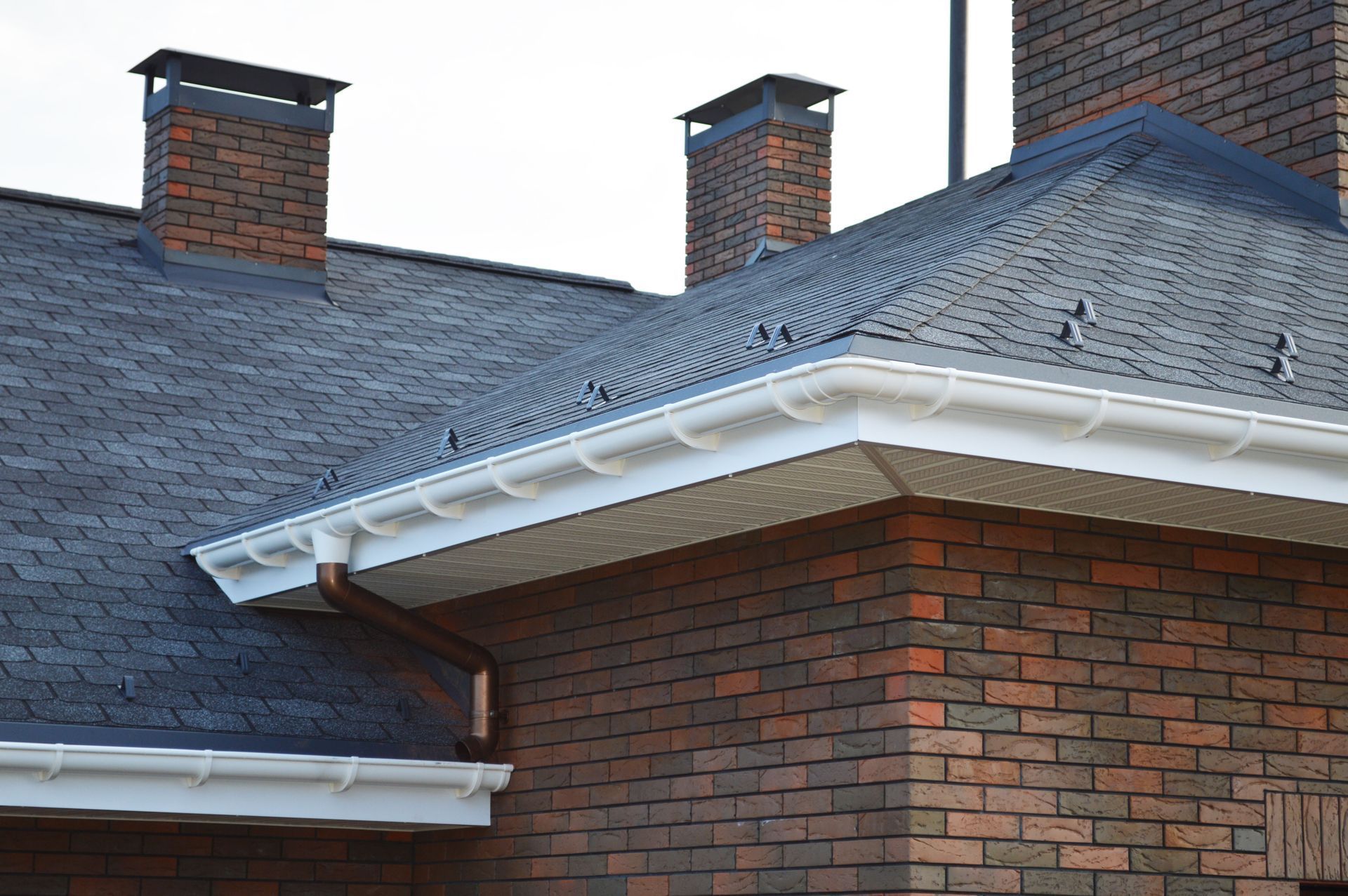 A close-up of a brick house roof with dark gray shingles, white gutters, two brick chimneys, and a copper downspout.