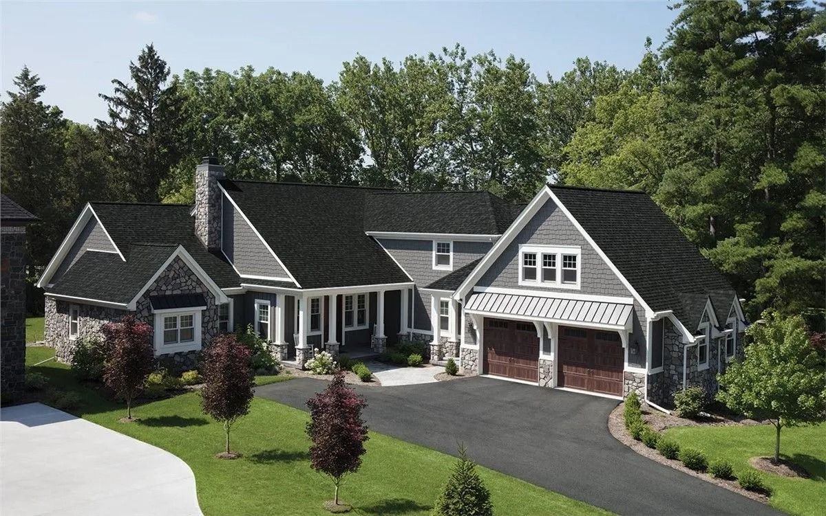 A grey shingled house with dark roofing and a stone facade, featuring a paved driveway and surrounded by trees.