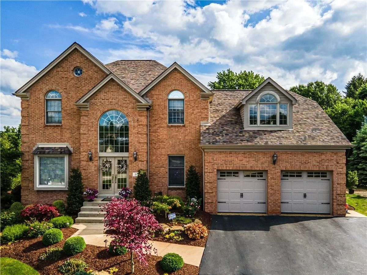 Two-story brick house with a two-car garage, arched entryway, and landscaped front yard under a blue sky with clouds.