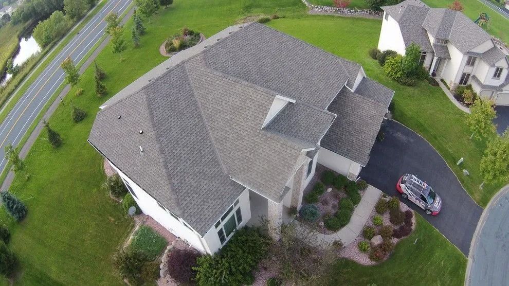 An aerial view of a suburban house with a gray shingled roof, a paved driveway with a parked van, and surrounding lawn.