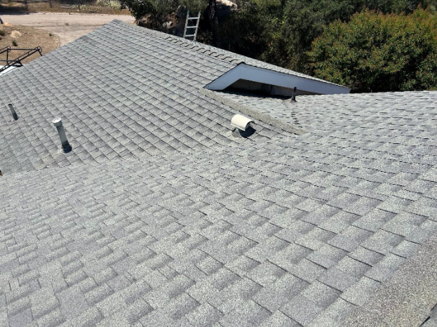 High-angle view of a grey shingled roof with a small dormer window, vent pipe, and chimney in a residential setting.
