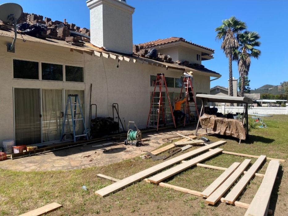 Workers on ladders perform roofing repairs on a stucco house with a patio containing tools and construction materials.