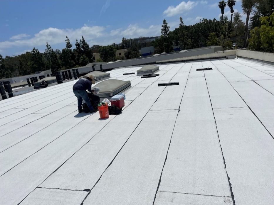 A worker crouches on a flat, white-coated commercial roof with equipment and supplies nearby.
