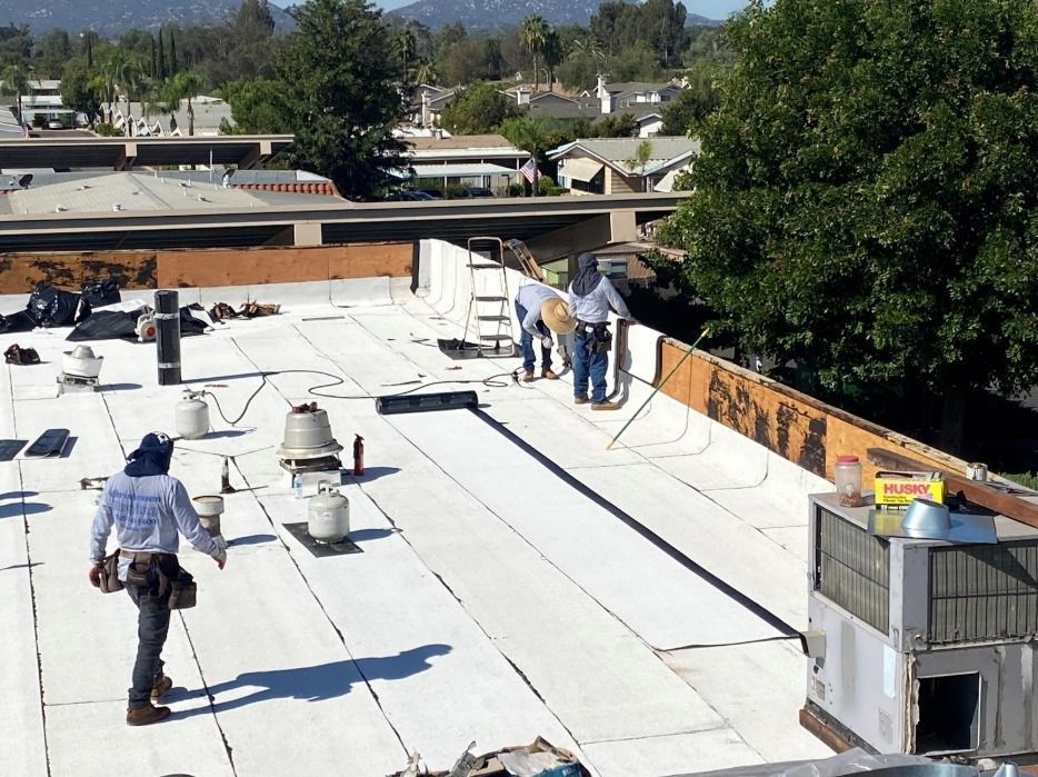 Workers in work clothes applying a white membrane roof coating on a building rooftop.