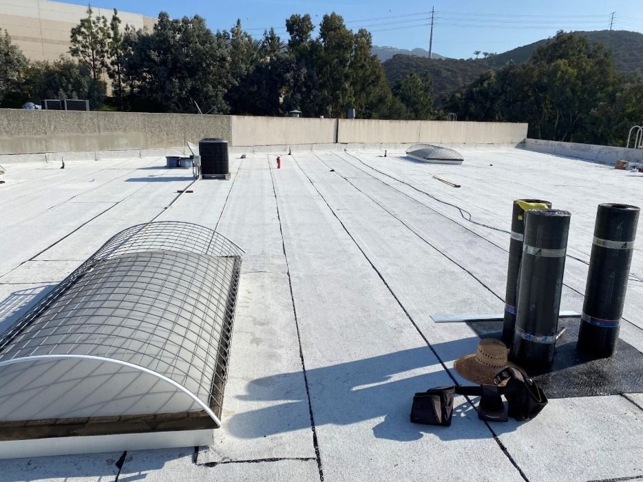 A flat white roof with a long skylight, ventilation pipes, and work gear under a bright blue sky.