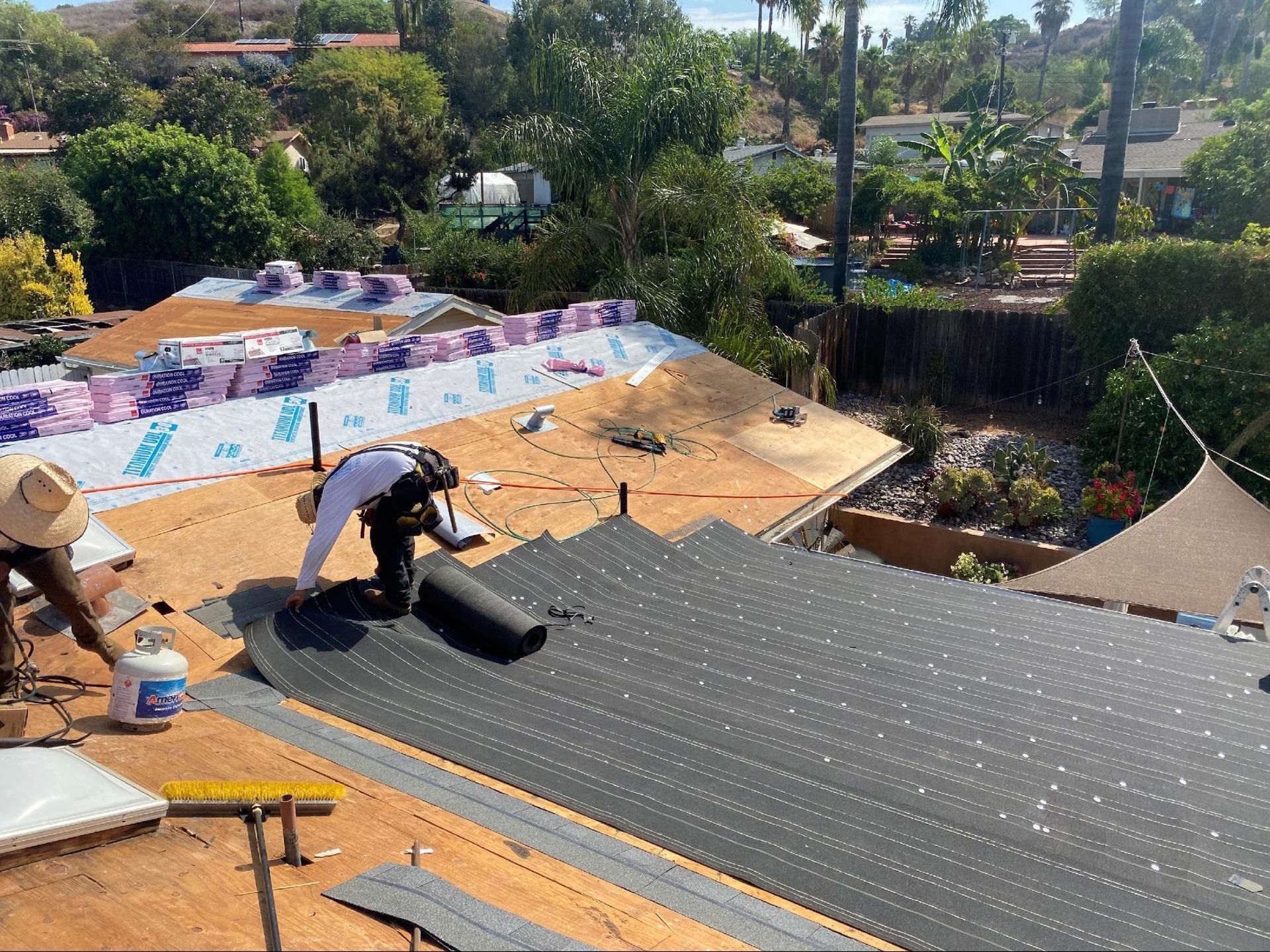 A construction worker installing black roofing underlayment on a residential roof in a sunny, suburban backyard setting.