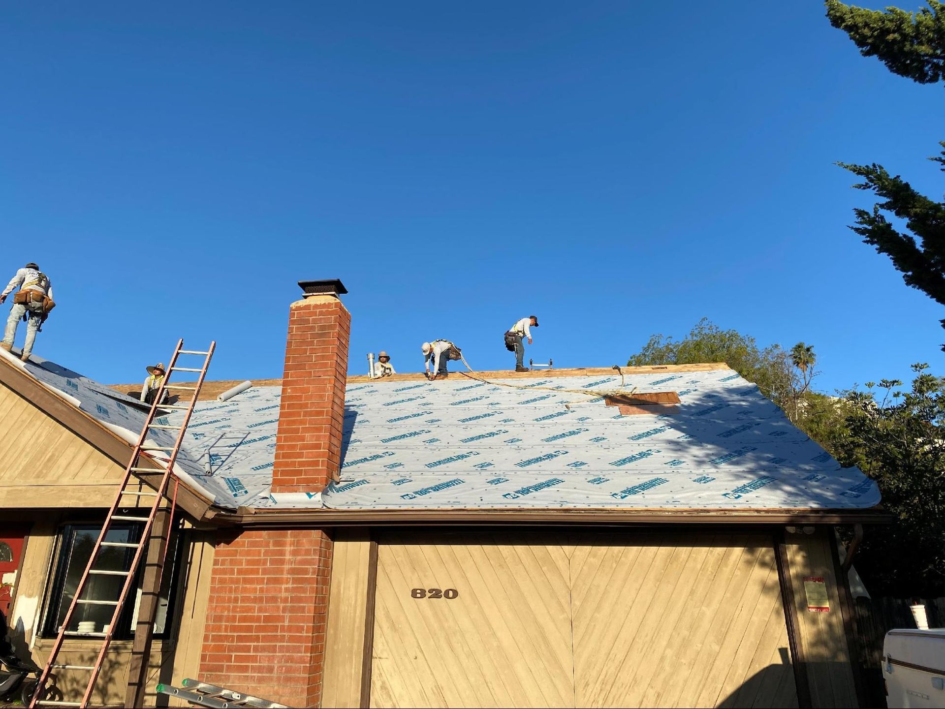 Several workers on a house roof covered in blue underlayment, with a brick chimney and a wooden garage door below.